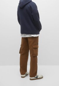 Person klædt i brune cargo-bukser, hvide sneakers med sorte striber, en hvid skjorte og en marineblå hoodie, der står foran en ensfarvet baggrund.