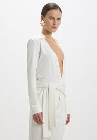 Blusa bianca a maniche lunghe con scollatura a V profonda e chiusura a bottoni, caratterizzata da un dettaglio in vita legato e un tessuto texturizzato. Orecchini circolari dorati.