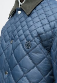 Steppjacke in Blau mit einem Cordkragen, die ein rundes Logo und einen Druckknopfverschluss aufweist. Das Material hat eine glatte, glänzende Oberfläche.