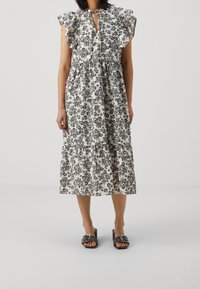 Krémové a černé midi šaty s květinovým vzorem, s volánkovými rukávy, šňůrkou u výstřihu a vrstvenou sukní. Doplňují se černými sandály na klínu.