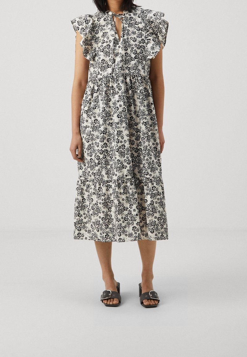 Krémové a černé midi šaty s květinovým vzorem, s volánkovými rukávy, šňůrkou u výstřihu a vrstvenou sukní. Doplňují se černými sandály na klínu.