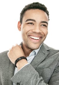 Lächelnder Mann in grauem Blazer, der ein schwarzes Perlenarmband an erhobener Hand zeigt, mit hellblauem Hemd und lockigem, kurzem Haar vor weißem Hintergrund.