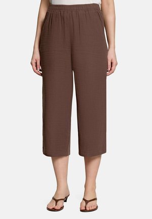 Bruine losse cropped broek met zijzakken gedragen door een staand persoon, gecombineerd met bruine sandalen met hak en teenslipper.