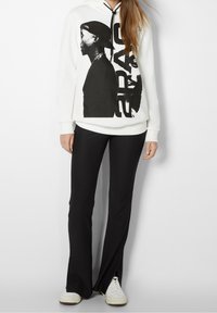 Sudadera blanca con estampado gráfico negro que presenta una silueta y texto. Combinada con pantalones ajustados negros y zapatillas blancas.