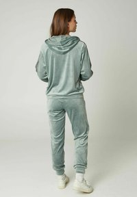 Samt-Trainingsanzug in gedämpftem Grün, bestehend aus einer Kapuzenjacke und taillierten Hosen. Weiches Material mit gerippten Bündchen und weißen Sneakern.