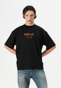 T-shirt in cotone nero con spalle scese, caratterizzato da testo e simboli rossi "REPLAY" sulla parte anteriore. Indossato con jeans blu strappati.