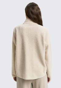 Haut en polaire beige avec une texture douce, dotée d'un col montant et de manches longues. Ourlet arrondi et coutures minimalistes pour une coupe décontractée.