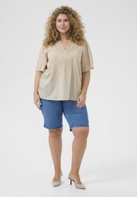 Beige blouse with short sleeves and vertical pleats at the neckline, paired with blue pinstriped shorts. Model wearing light-colored heels.