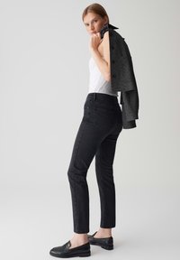 Jeans skinny neri con vita alta e una leggera elasticità, abbinati a una canotta bianca e mocassini neri. Un blazer scuro e strutturato è appoggiato sulla spalla.
