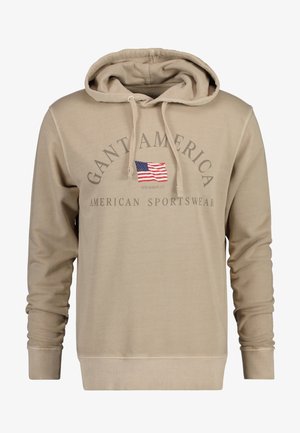 Sweat à capuche beige avec cordons, arborant un petit drapeau américain et les inscriptions « GANT AMERICA » et « AMERICAN SPORTSWEAR » sur le devant.