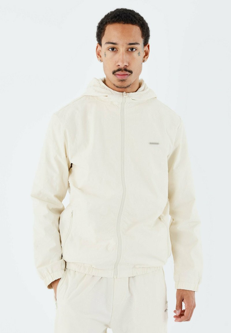 Veste zippée beige clair avec capuche ; présente des poignets et un ourlet élastiques, une texture de motif subtile et des poches latérales. Détail du logo de marque réfléchissant.