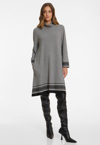 Schwarz-weiß kariertes Kleid mit hohem Kragen, langen Ärmeln, Seitentaschen und dekorativem gemustertem Saum, kombiniert mit knielangen schwarzen Stiefeln.