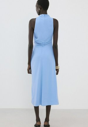 Personne portant une robe midi sans manches bleu clair, debout de dos sur un fond uni, accessoirisée avec des bracelets blancs et des chaussures noires.