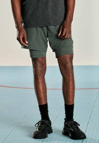 Herreshorts i mørkegrøn, letvægtsmateriale. Har et lagdelt design og er parret med sorte sokker og sneakers på et struktureret gulv.