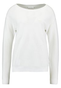 Vit långärmad tröja med ribbat mönster, rund halsringning och en liten Lacoste-logotyp på bröstet. Mjuk material och avslappnad passform.