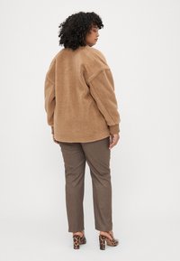 Veste en polaire beige avec une coupe ample, des manches larges et des poignets côtelés, associée à un pantalon marron et des chaussures à talons carrés à motifs.