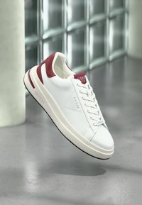 Hvit lær sneakers med røde semskede detaljer, teksturerte lisser og gummi såle. Har "GUESS" merkevare på siden og tungen.