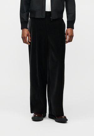 Homme portant un pantalon ample en velours noir, des chaussures marron foncé et une veste zippée noire avec une chemise blanche en dessous.
