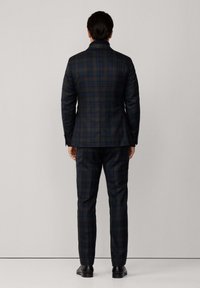 Uomo in piedi, rivolto di spalle, che indossa un abito a quadri scuri con pantaloni su misura e scarpe lucide nere su uno sfondo semplice.