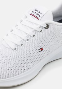 Białe buty sportowe z teksturowaną siateczkową cholewką, płaskimi białymi sznurowadłami, okrągłym metalowym oczkiem oraz małym logo w kolorach czerwonym, białym i niebieskim na boku.