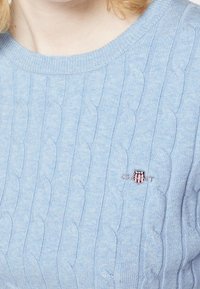 Pull en maille torsadée bleu clair avec un petit emblème en forme de bouclier rouge, blanc et bleu sur la poitrine, porté par une personne à la peau claire et aux cheveux clairs.