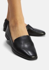 Scarpe slip-on in pelle nera indossate a piedi nudi con tacco basso su uno sfondo bianco, mostrando le viste frontale e laterale.