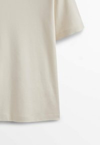 T-shirt beige en coton à manches courtes suspendu sur un fond blanc, montrant les détails du milieu et des manches.