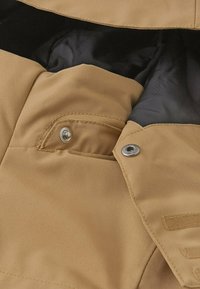 Beige Stoffjacke mit glatter Textur, metallischen Druckknopfverschlüssen und einem schwarzen Innenfutter. Enthält Taschen und gestickte Akzente.