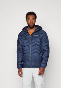Doudoune marine avec capuche, présentant un motif matelassé en chevrons et une fermeture éclair. Portée par-dessus une chemise orange, avec un pantalon blanc visible.