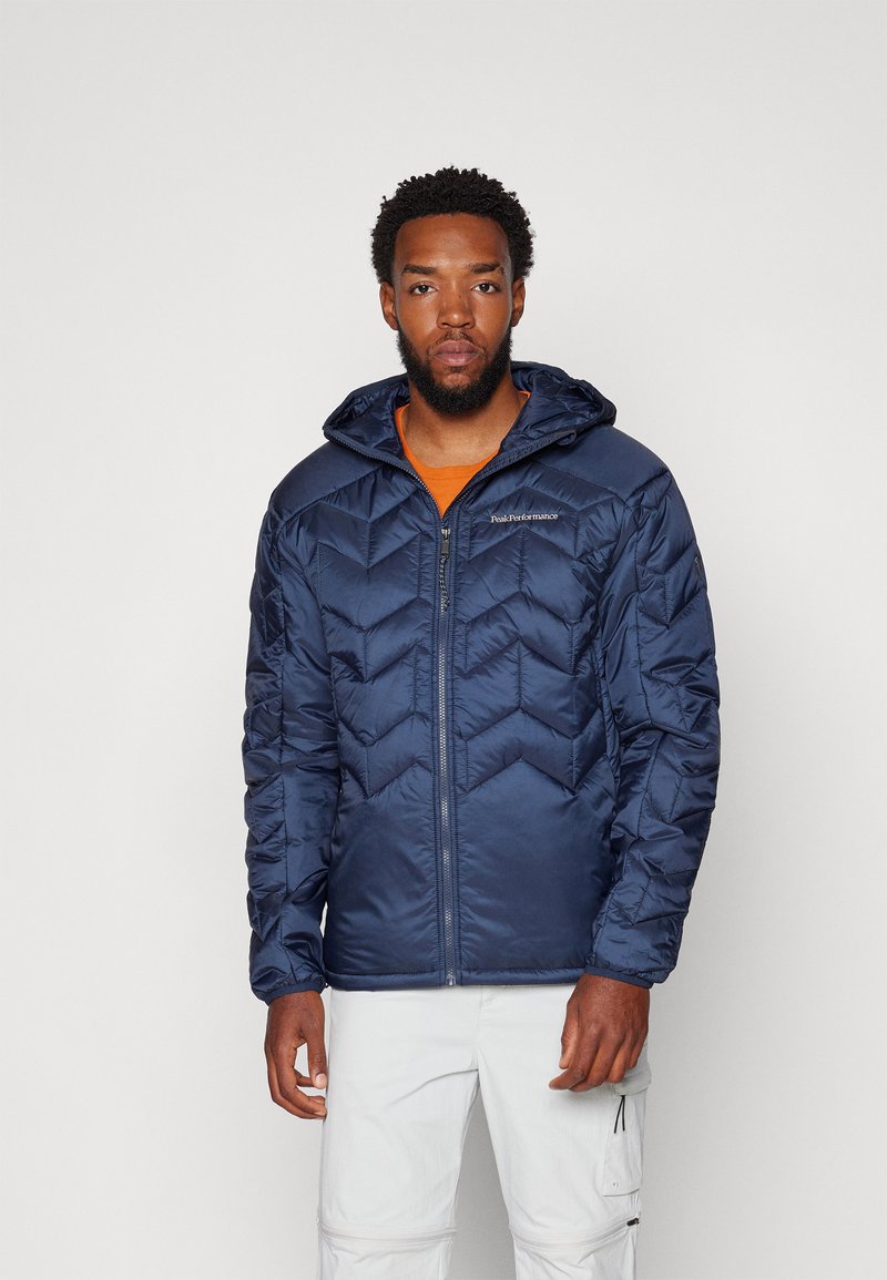 Doudoune marine avec capuche, présentant un motif matelassé en chevrons et une fermeture éclair. Portée par-dessus une chemise orange, avec un pantalon blanc visible.