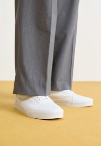 Baskets en toile blanches avec semelles en caoutchouc plates, associées à un pantalon gris bien taillé. La texture est lisse et la coupe semble décontractée.