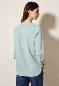 Blusa azzurra chiara, vestibilità ampia con maniche a tre quarti, un yoke posteriore e dettagli a pieghe. Abbinata a pantaloni blu navy con righe sottili.