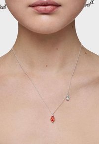 Collar de cadena de plata con un colgante de gema roja ovalada y un pequeño acento de plata, sobre un fondo de piel de tono neutro.