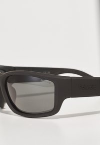 Czarne okulary przeciwsłoneczne Polaroid o matowym wykończeniu, z ciemnymi przyciemnionymi soczewkami oraz geometrycznym designem oprawki. Wyraźne logo na skroni.