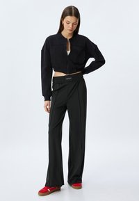 Zwarte cropped zip-up jack met een ronde hals, gecombineerd met hoge, brede zwarte broeken met een gerimpelde tailleband en rode sneakers.