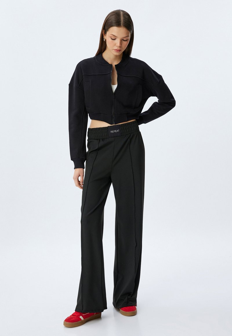 Zwarte cropped zip-up jack met een ronde hals, gecombineerd met hoge, brede zwarte broeken met een gerimpelde tailleband en rode sneakers.