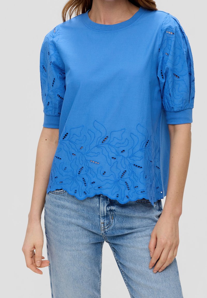 Femme portant un chemisier bleu à manches courtes avec des découpes brodées et des manches bouffantes, assorti à un jean bleu clair.