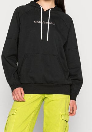 Persoon draagt een zwarte Converse hoodie met witte trekkoorden en felgele cargobroek, staand tegen een effen achtergrond.