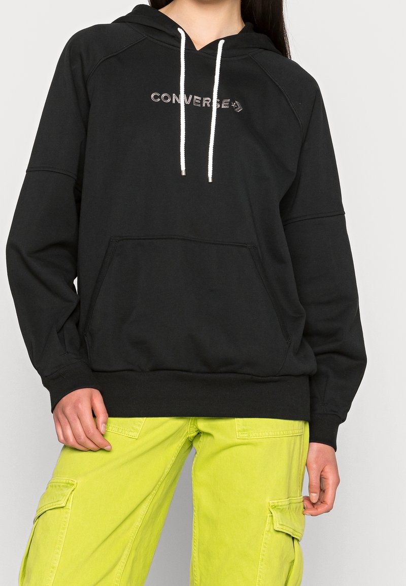 Personne portant un sweat à capuche Converse noir avec des cordons blancs et un pantalon cargo jaune vif, debout devant un fond uni.