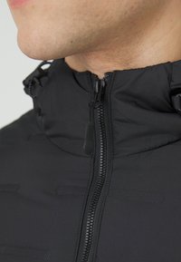 Giacca nera con colletto alto, zip frontale e cordini regolabili. Tessuto liscio con accenti di cuciture orizzontali.