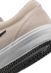 Zapatilla de ante beige con una media suela blanca y una suela exterior de goma negra texturizada. Presenta un logo de Nike negro en contraste y detalles de costura visibles.
