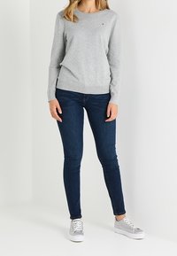 Svetle šedý svetr s kulatým výstřihem a dlouhými rukávy, nošený s tmavě modrými úzkými džíny. Model má na sobě šedé tenisky s bílými podrážkami.