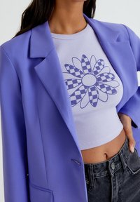 Donna che indossa un top cropped lilla con un motivo a fiore a scacchi, abbinato a un blazer viola e a jeans a vita alta grigio scuro.