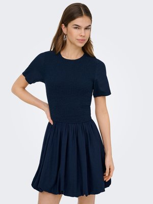 Kvinne iført en kortermet marineblå kjole med rynket livstykke og samlede folder i skjørtet, stående med en hånd på hoften.