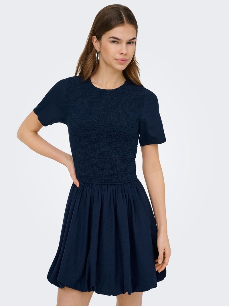 Naine, kes kannab tumesinist lühikeste varrukatega kleiti, millel on tikitud ülakeha ja kogutud seelik, seisab ühe käega puusal.