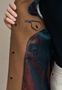 Bruine wollen overjas met een interieur dat een teal- en bordeauxrood patroon heeft, met een zwart label waarop "NEXT SIGNATURE" staat, maat M.
