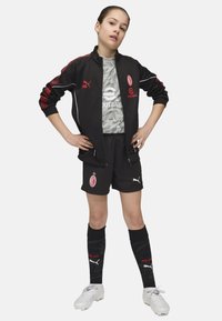 Chaqueta deportiva negra con acentos rojos, camiseta de camuflaje gris, pantalones cortos negros, calcetas negras con detalles rojos y botas de fútbol blancas.