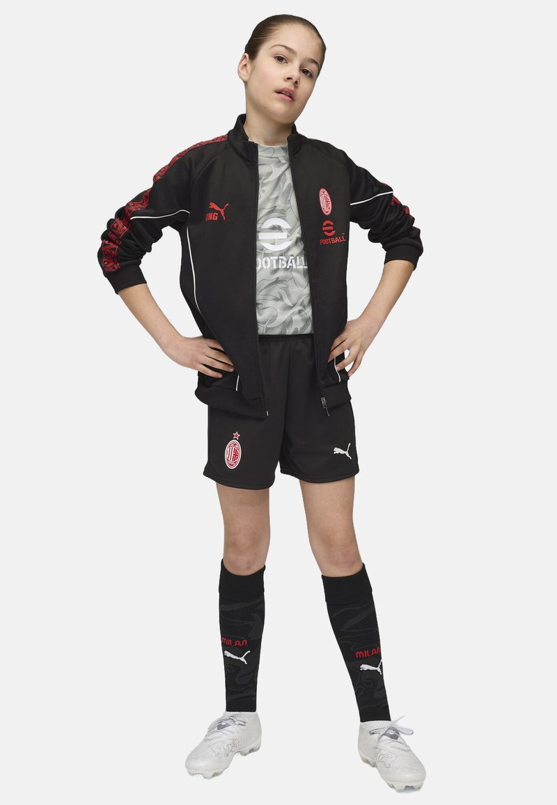 Chaqueta deportiva negra con acentos rojos, camiseta de camuflaje gris, pantalones cortos negros, calcetas negras con detalles rojos y botas de fútbol blancas.