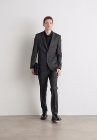 Grijs op maat gemaakte pak met een enkele knoop, zwarte overhemd eronder en een gladde textuur. Model houdt een zwarte clutch vast en draagt zwarte schoenen.