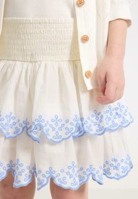 Hvid smocket skirt med lagdelte volanter og blå blomstret broderi langs kanten. Akkompagneret af en lysefarvet cardigan med træknapper.